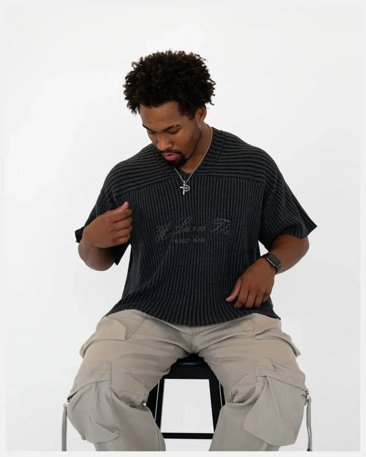 Man wearing black ribbed Christian graphic t-shirt and beige cargo pants, seated on stool