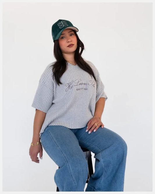 Woman in casual luxury apparel, ribbed gray shirt, blue jeans, green 99 cap, seated on stool