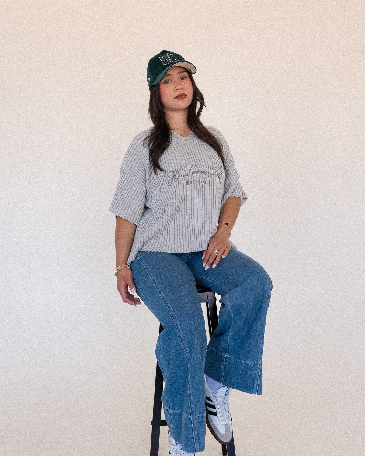 Woman in a grey ribbed t-shirt, blue wide-leg jeans, and a dark green baseball cap sitting on a stool.