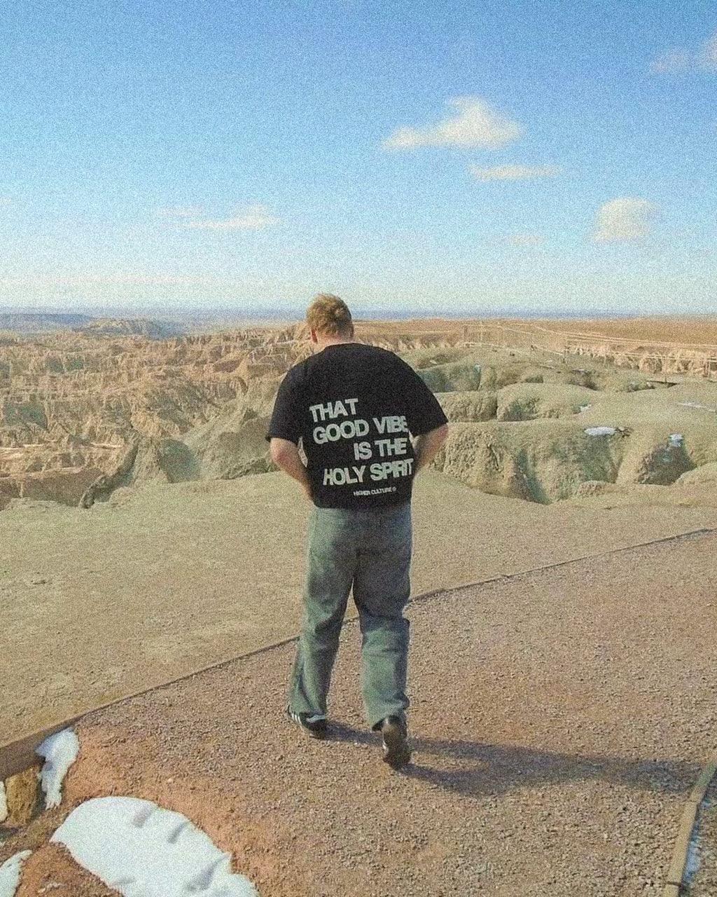 Man in black t-shirt with 'That good vibe is the Holy Spirit' text overlooking a canyon.