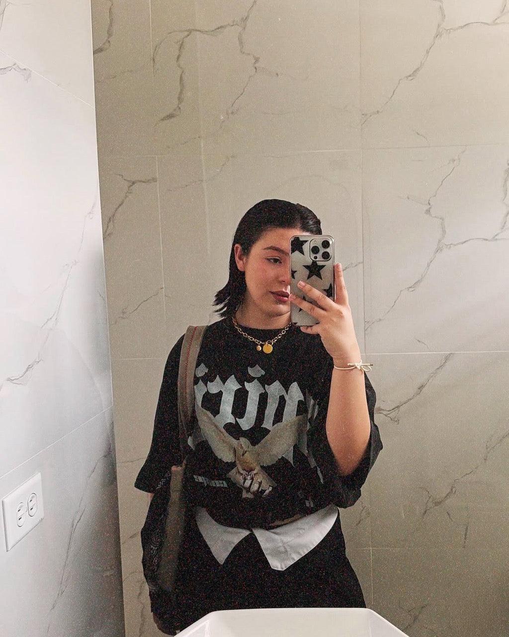 Woman in a black graphic t-shirt with white print taking a mirror selfie in a marble bathroom.